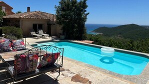 A pool area with a view of the sea, outdoor furniture, and a building with a tiled roof.