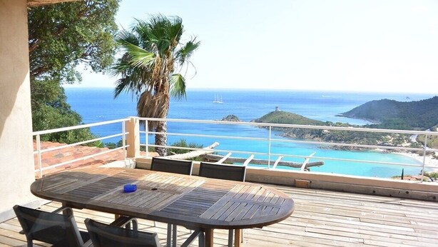 A wooden deck with a table and chairs, overlooking a coastal view with a clear sky, a palm tree, and a distant island.