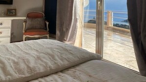 A bedroom with a bed, a chair, a desk, and a painting. There is a sliding glass door leading to a balcony with a view of the sea.