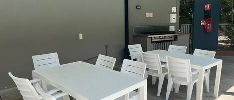 An outdoor patio with white plastic tables and chairs, a barbecue grill, and a fire extinguisher.