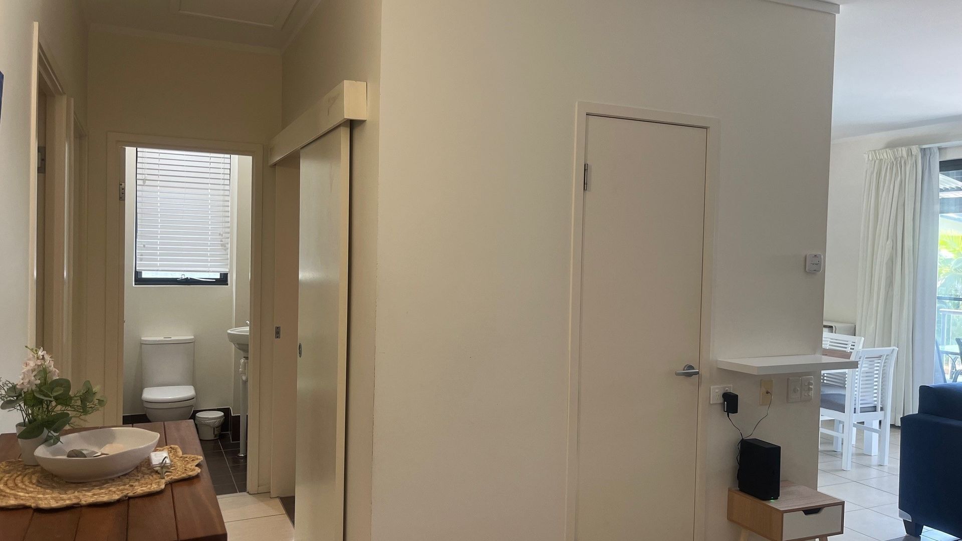 A hallway leading to a bathroom with a toilet, sink, and window with blinds. There is a wooden table with a bowl and a plant.
