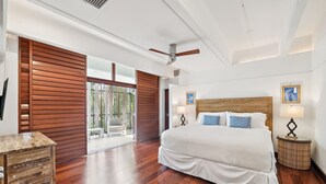 Une chambre spacieuse avec un grand lit, des stores en bois, un ventilateur de plafond et une vue sur l’extérieur grâce à une porte vitrée.