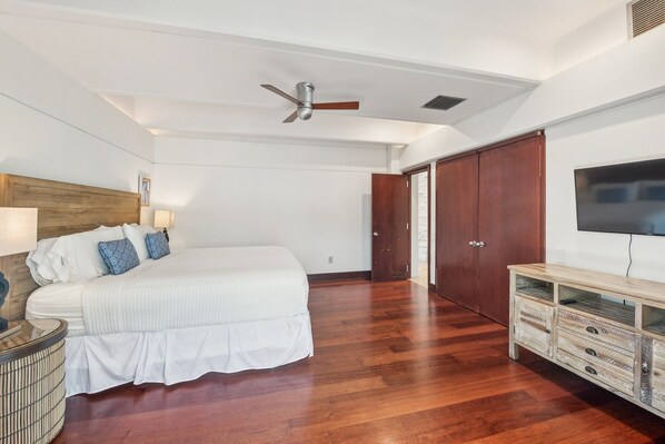 A bedroom with a bed, a ceiling fan, a TV, and wooden flooring.