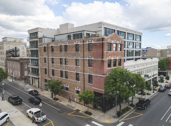 Un edificio de varios pisos con una combinación de fachadas de ladrillo y paneles de vidrio modernos, situado en una esquina con marcas viales visibles y coches estacionados.