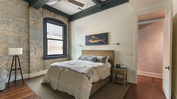 A bedroom with a brick wall, a wooden ceiling, a bed with a headboard, a nightstand, a lamp, and a window.