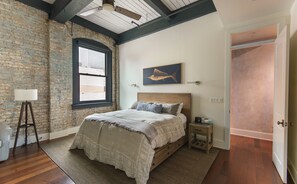 A bedroom with a brick wall, a wooden ceiling, a bed with a headboard, a nightstand, a lamp, and a window.
