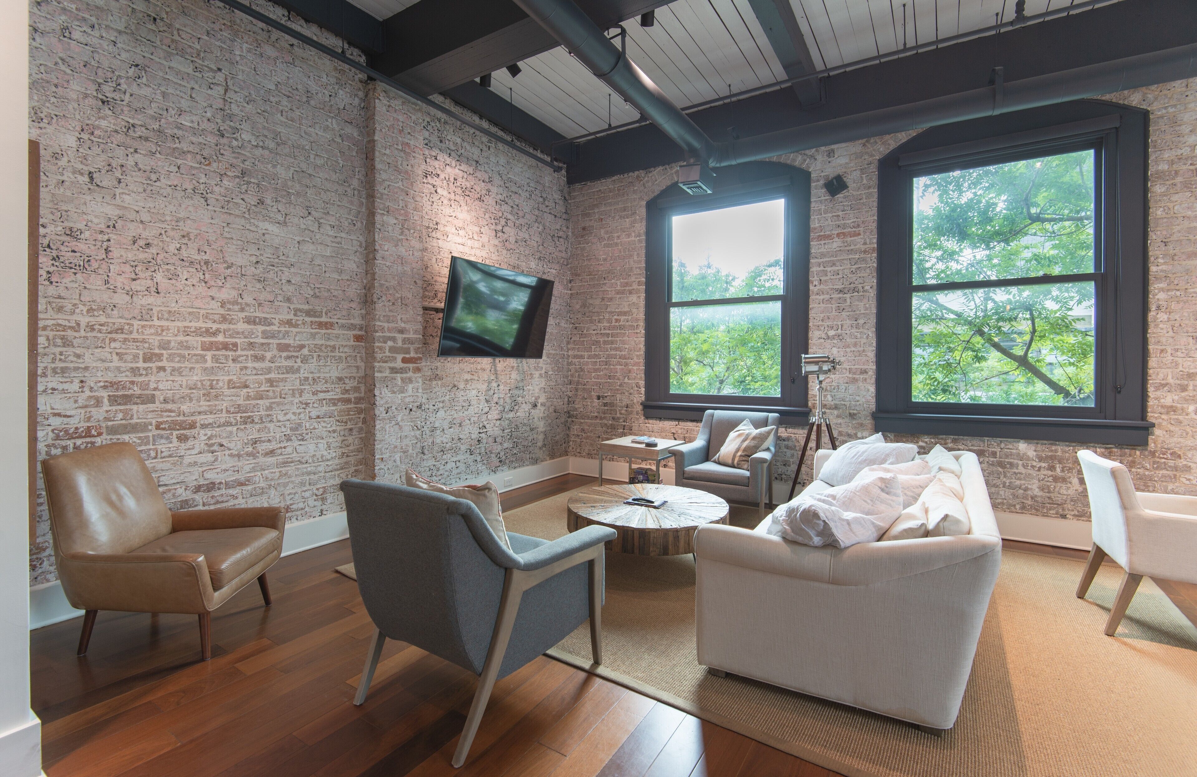 A spacious room with a brick wall, a flat-screen TV mounted on it, and large windows providing a view of trees.