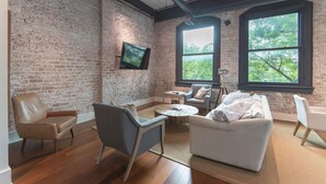 A spacious room with a brick wall, a flat-screen TV mounted on it, and large windows providing a view of trees.