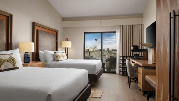 A hotel room with two beds, a desk, a chair, a TV, and a balcony with a view of a tower.