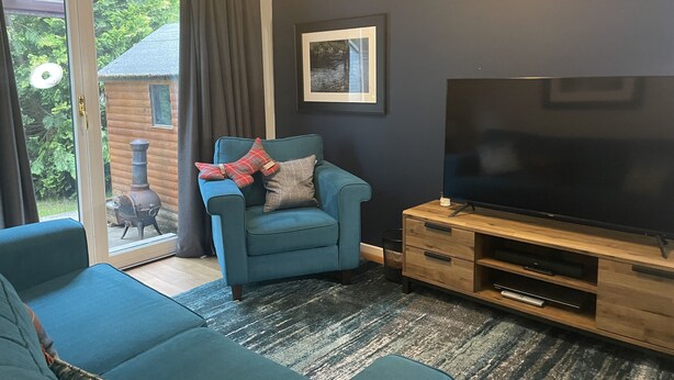 Ein Wohnzimmer mit einem blauen Sofa, einem Fernseher auf einem Holzschrank und einem großen Fenster mit Vorhang.