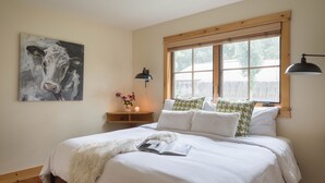 Un dormitorio con una cama grande, una ventana con persianas, una lámpara de noche, una lámpara de pared y un cuadro de una vaca.