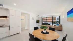 A modern living room with a round wooden table, black chairs, a sofa, and a large window offering a view of a building. - 405 PINE TREE | Short walk to beach | Accom Holidays (TERRIGAL)