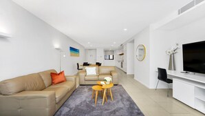 A modern living room with a sofa, a coffee table, and a TV. There is a dining area in the background. - 405 PINE TREE | Short walk to beach | Accom Holidays (TERRIGAL)