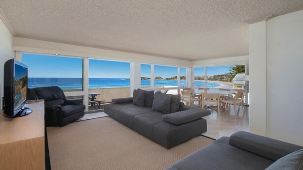 A modern living room with a large gray sofa, a flat-screen TV, and a dining area with wooden chairs. - MANYANA | Accom Holidays (WAMBERAL)
