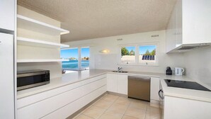 A modern kitchen with white cabinetry, a built-in oven, a dishwasher, and a window overlooking a scenic view. - MANYANA | Accom Holidays (WAMBERAL)
