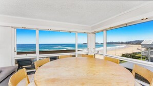 A room with a large table and chairs, a view of the beach, and a white ceiling. - MANYANA | Accom Holidays (WAMBERAL)