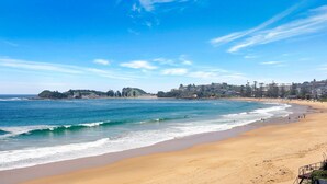 A beach with a clear view of the ocean, sandy shore, and a coastal town in the background. - MANYANA | Accom Holidays (WAMBERAL)