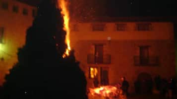 Un edificio con varias ventanas y balcones, un fuego ardiendo al frente y algunas personas en el fondo.