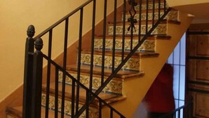 A staircase with a wooden handrail and black metal balusters, leading to a landing with a decorative wall and a light fixture. - LILA-ROOM-Rural House Angelita (Olocau Del Rey)