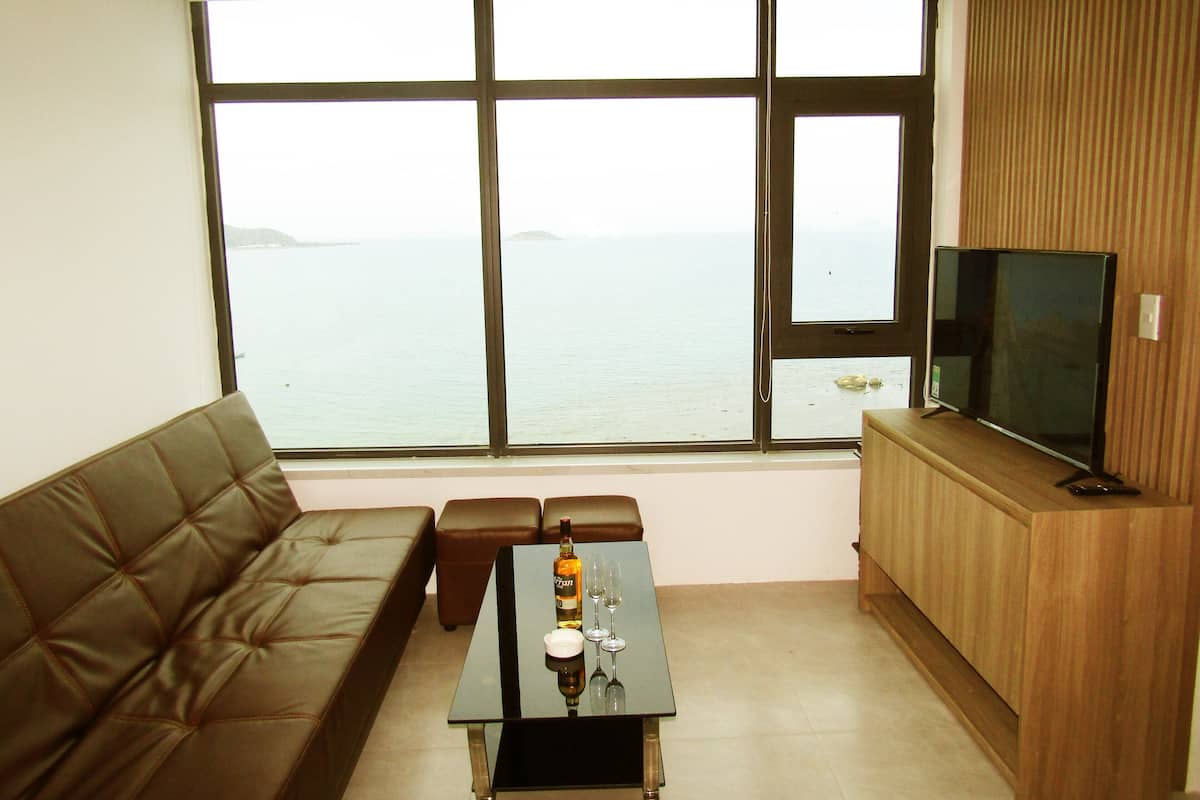 A living room with a large window, a brown leather sofa, a glass coffee table with a bottle and wine glass, and a TV on a wooden stand.