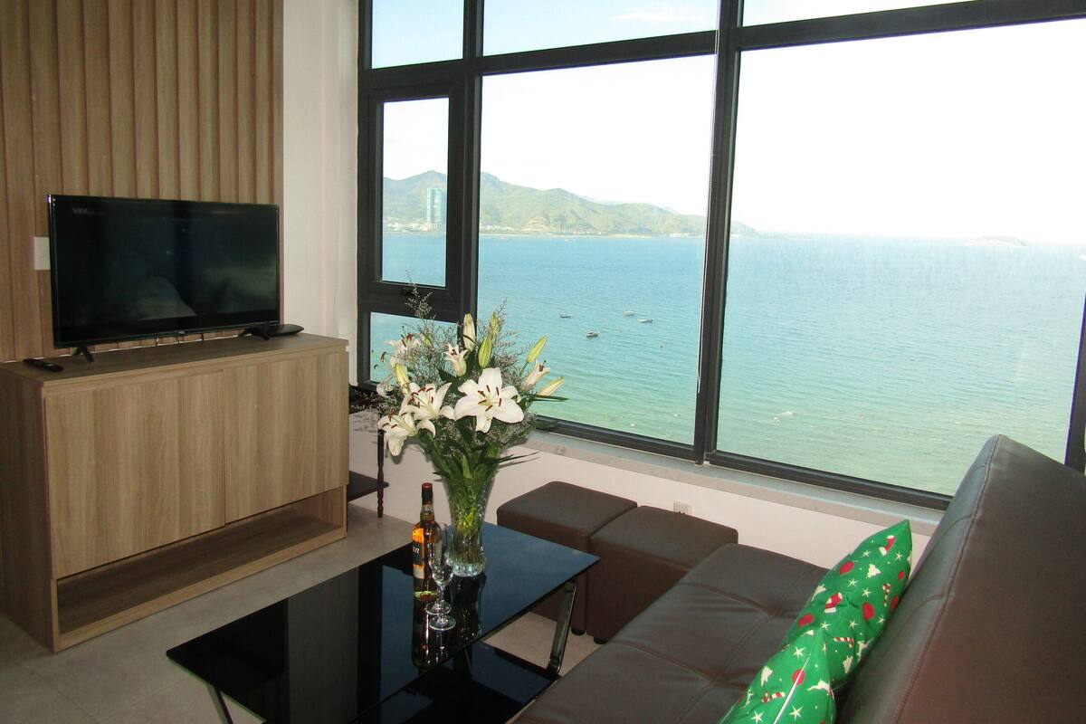 A modern living room with a large window, a flat-screen TV, a sofa, and a table with a vase of flowers.