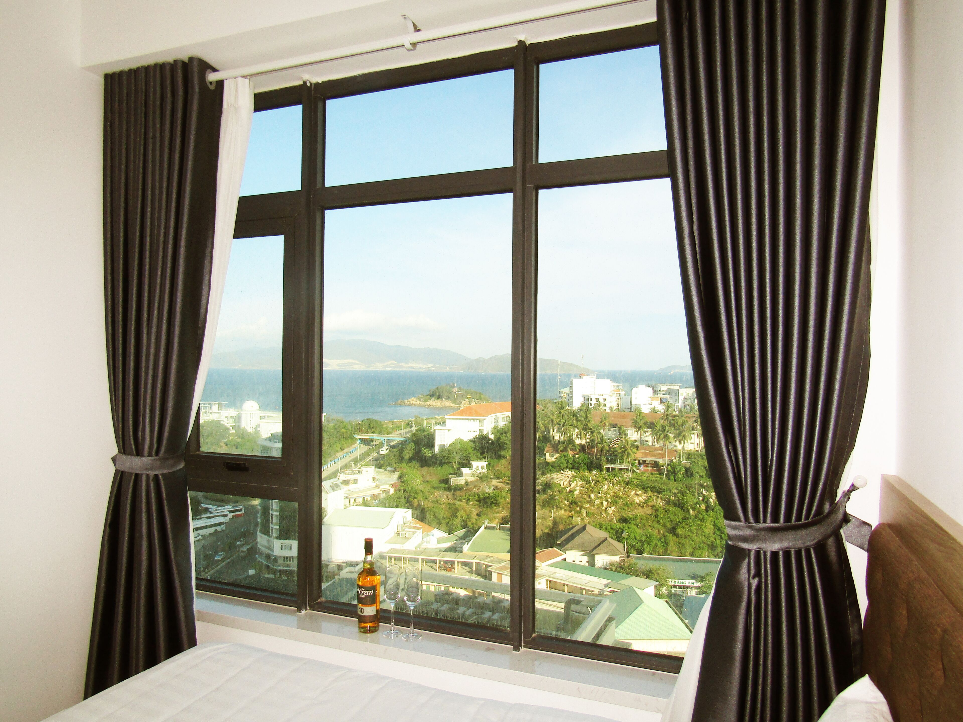 A hotel room with a large window, a bed, and a bottle of alcohol on the windowsill.