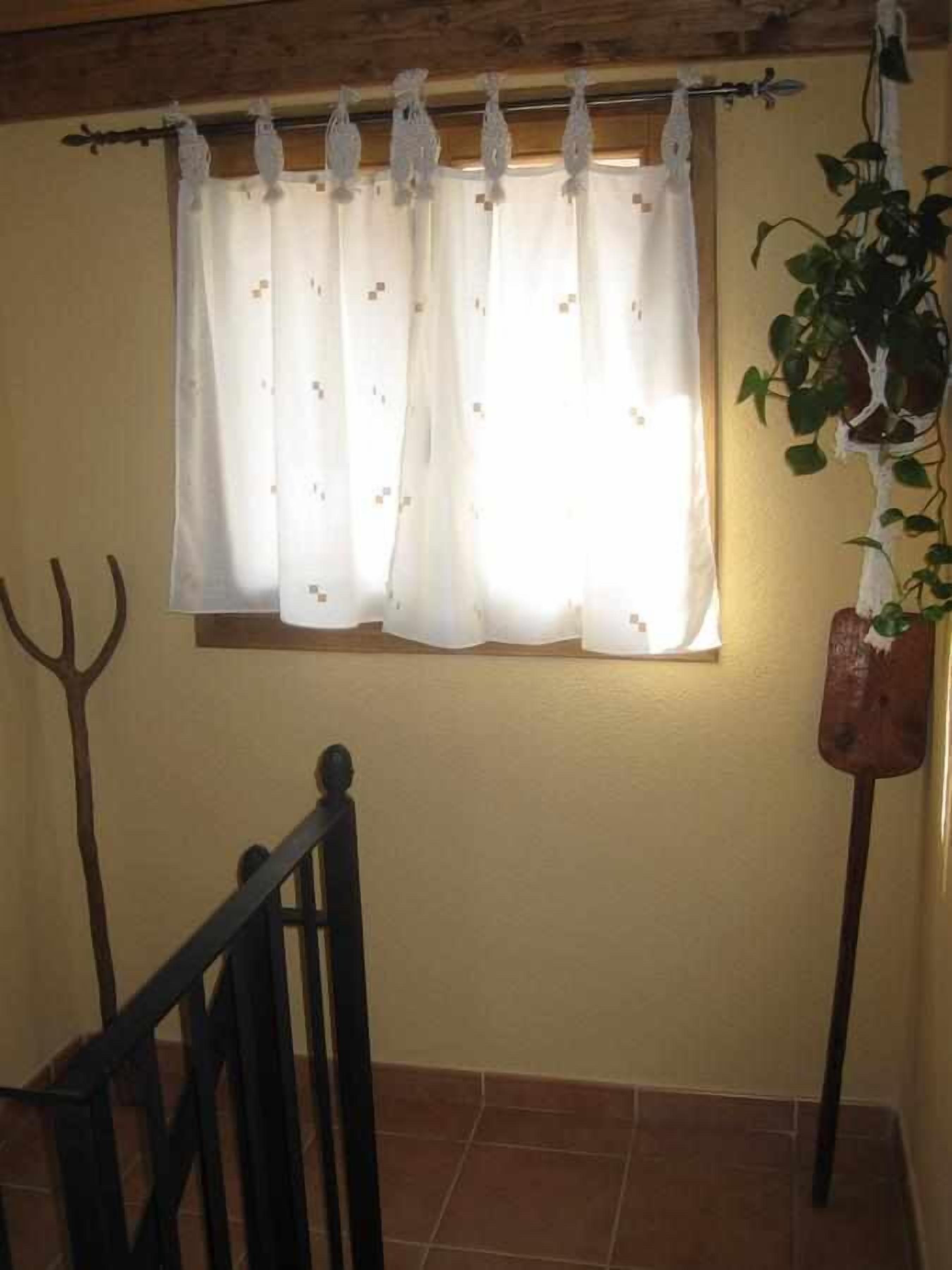 Una habitación con ventana cubierta por una cortina blanca con motivos florales, barandal de madera y planta en maceta a la derecha.
