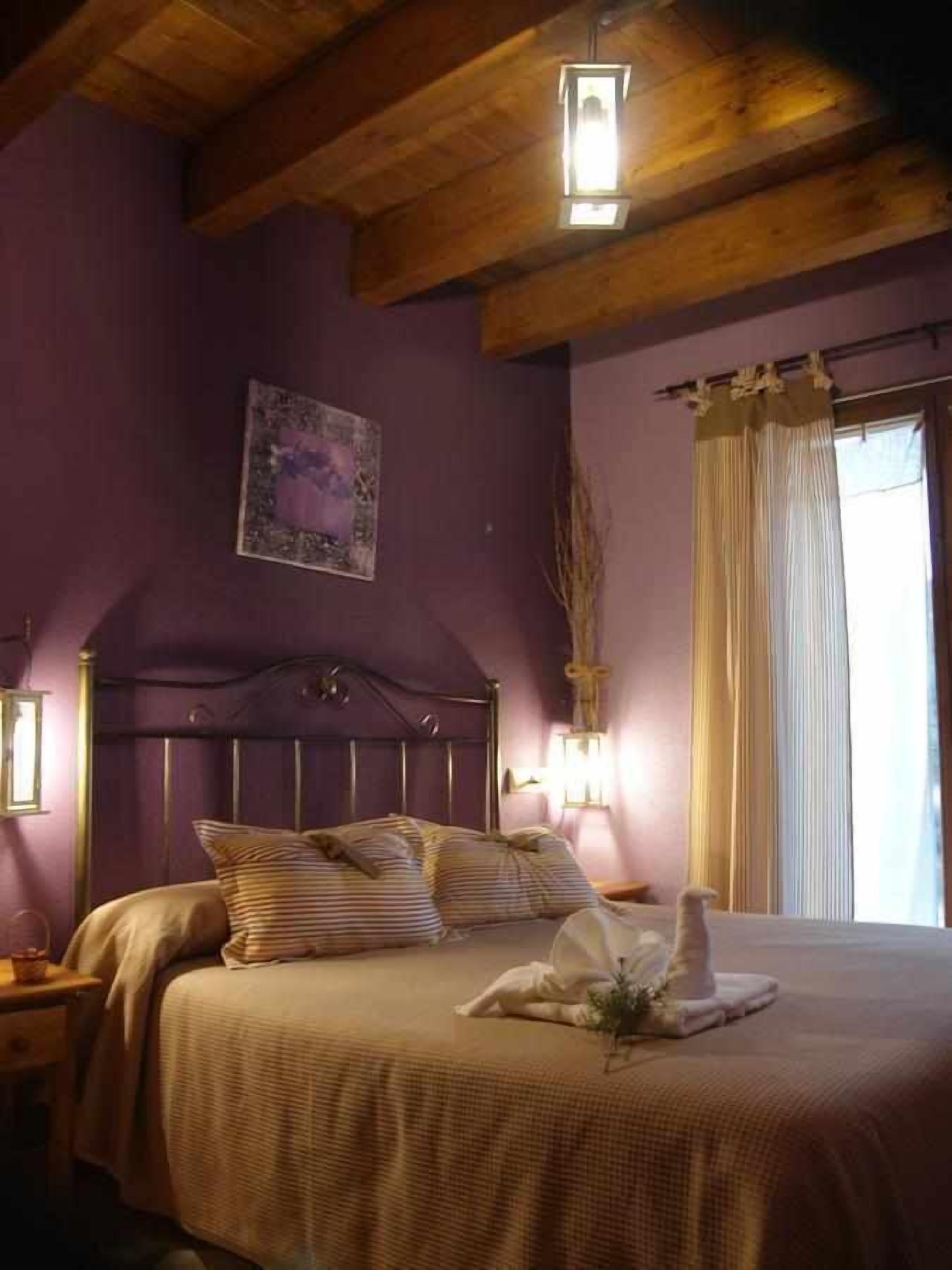 A bedroom with a wooden ceiling, a bed with striped bedding, a nightstand, a wall-mounted light, and a window with curtains.