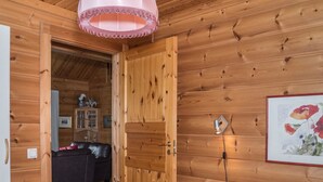 A wooden cabin interior with a bed, a door leading to a living area, and a framed artwork on the wall.