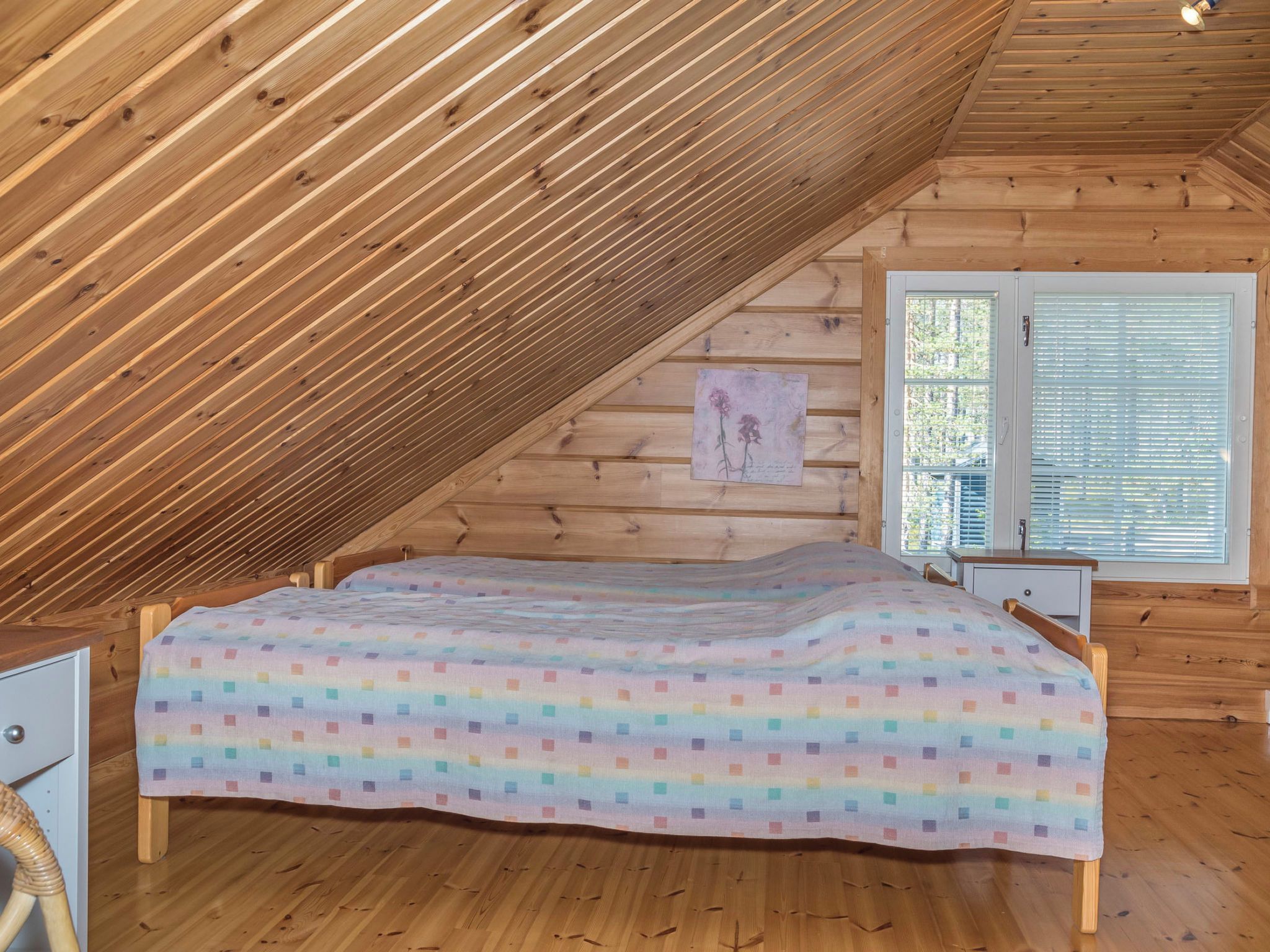 Un dormitorio con techo inclinado, una cama con colcha colorida, una mesita de noche, una silla y una ventana con persianas.