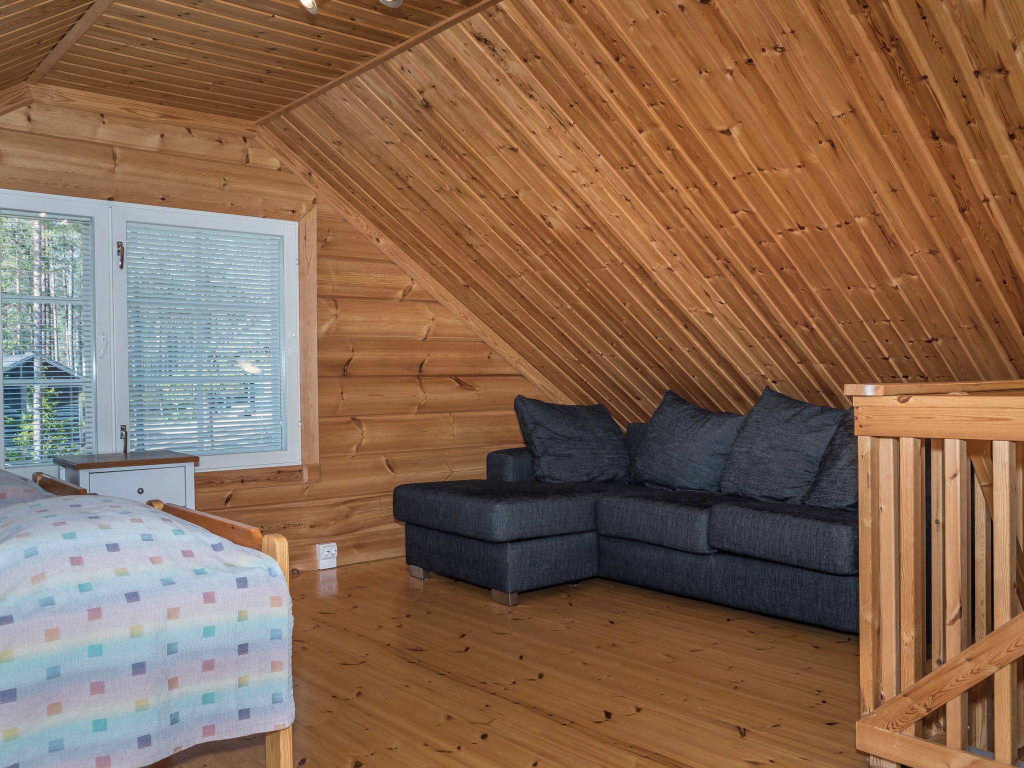 Una habitación acogedora con techo de madera, una cama con manta colorida, un sofá oscuro con cojines y una ventana con persianas.