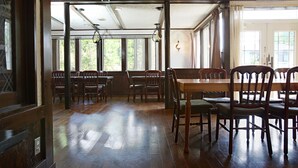 Een ruime eetruimte met houten tafels en stoelen, grote ramen en een plafond met zichtbare balken.