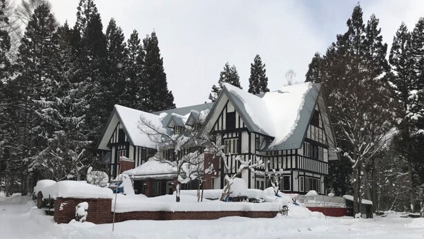 Een groot, meerlaags huis met een steil, met sneeuw bedekt dak, omgeven door hoge altijdgroene bomen.