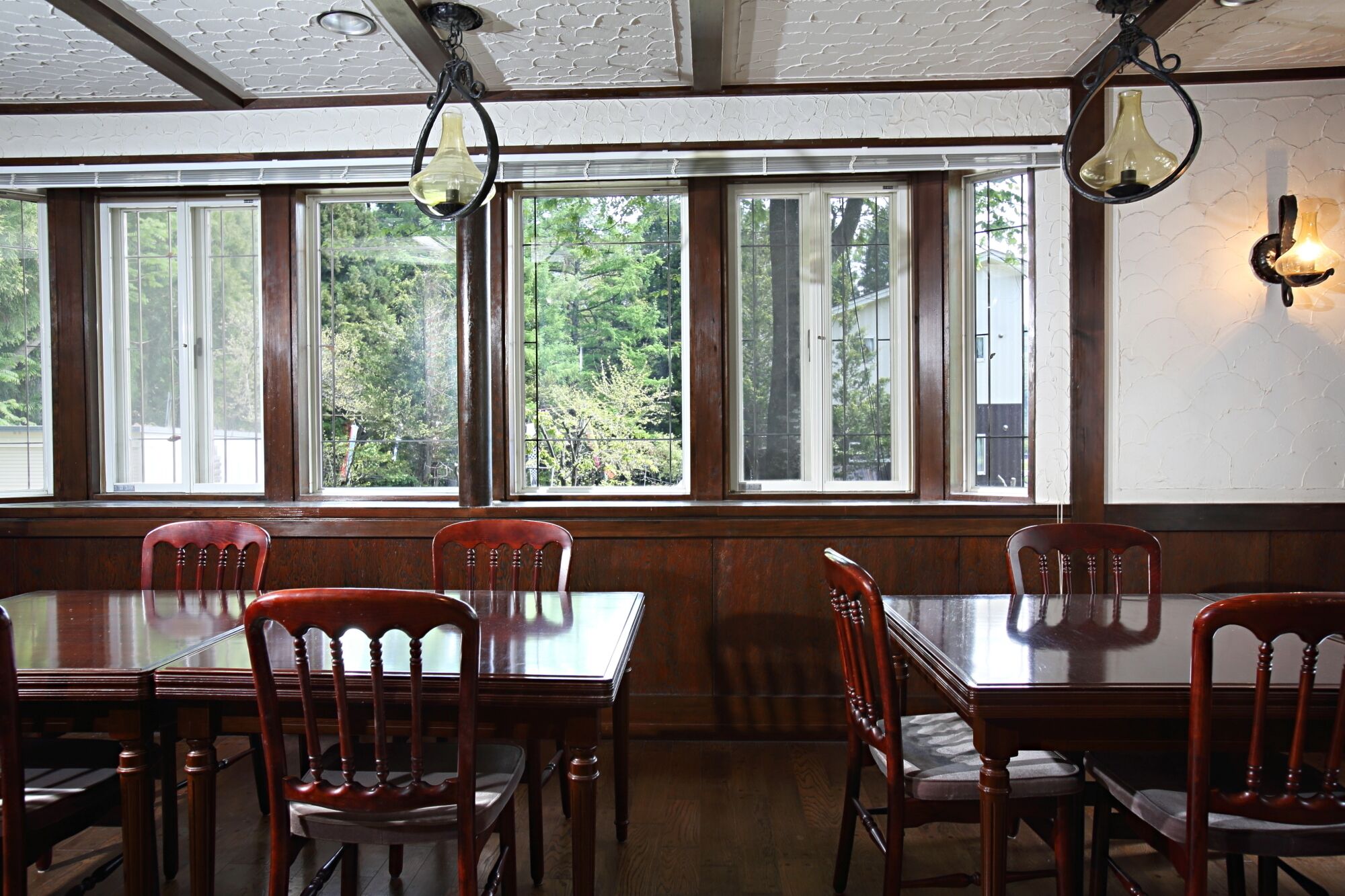 A dining area with wooden tables and chairs, large windows with a view of trees, and pendant lighting.