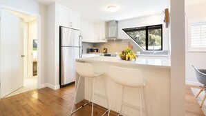 A modern kitchen with white cabinetry, a stainless steel refrigerator, a microwave, and a fruit bowl on the island. There are two white bar stools and a window with a view of greenery. - Luxury 3BR/2BA-1700 sq ft, Mountain view,BBQ (Whistler)