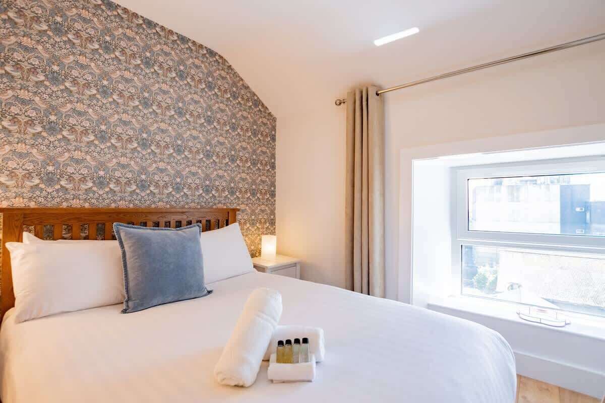 Una cama bien hecha con sábanas blancas y un almohéligo azul, un cabecero de madera, un papel tapiz estampado, una mesita de noche con lámpara y una ventana con cortinas.