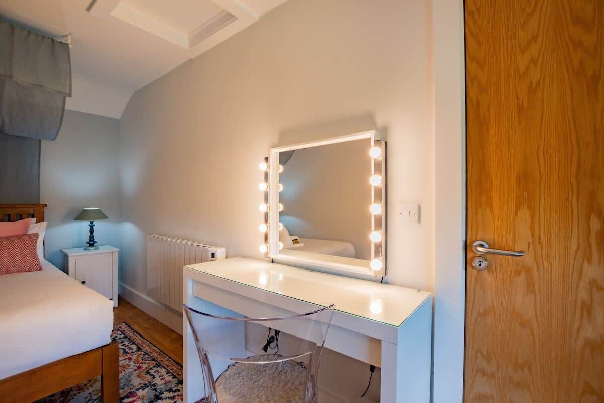 A bedroom with a bed, bedside table, lamp, vanity with a mirror and lights, and a wooden door.