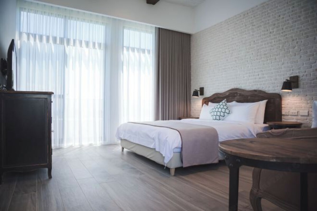 Un dormitorio con una cama, una mesita de noche de madera, un aparador y un ventanal con cortinas de gasa.