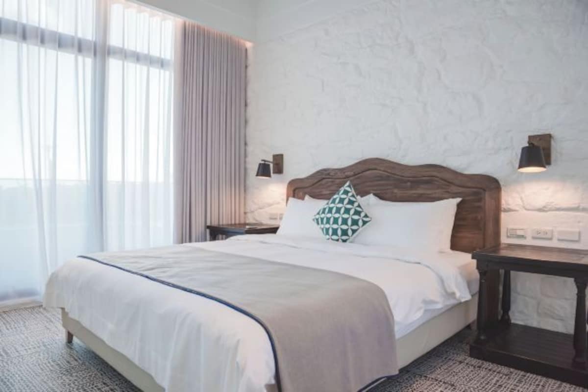 Una cama bien hecha con cabecera de madera, cubrecama blanco y almohada estampada en verde. Hay dos mesitas de noche con lámparas, y un ventanal con cortinas translúcidas.