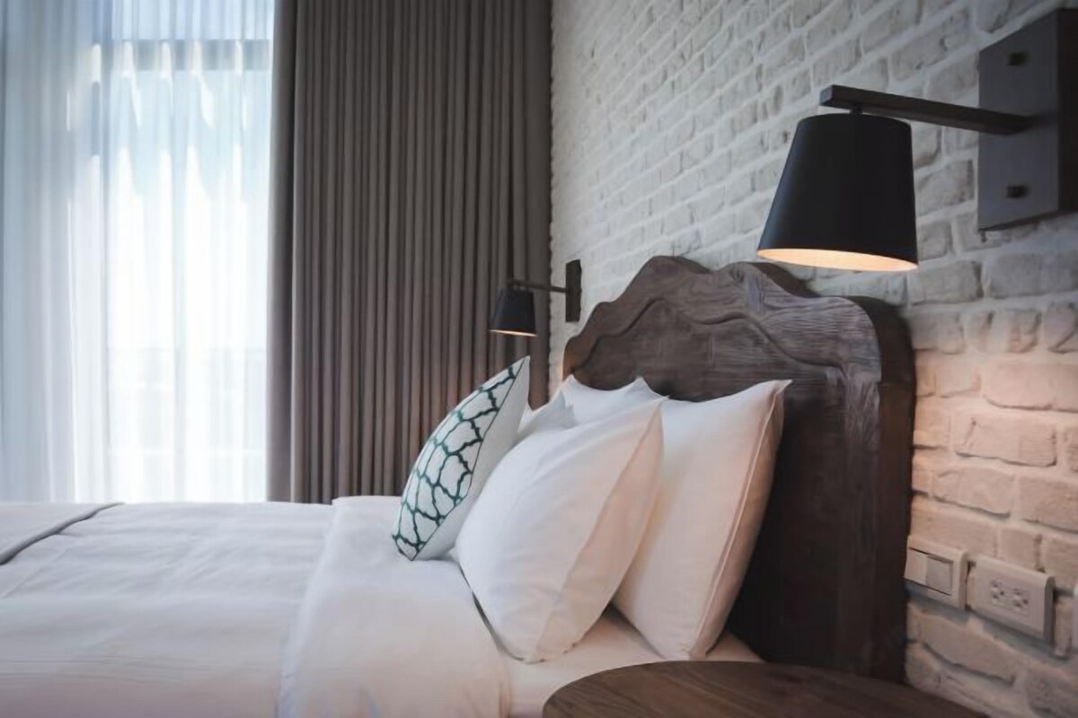 A hotel room with a bed, a wooden headboard, a wall-mounted lamp, and a window with curtains.