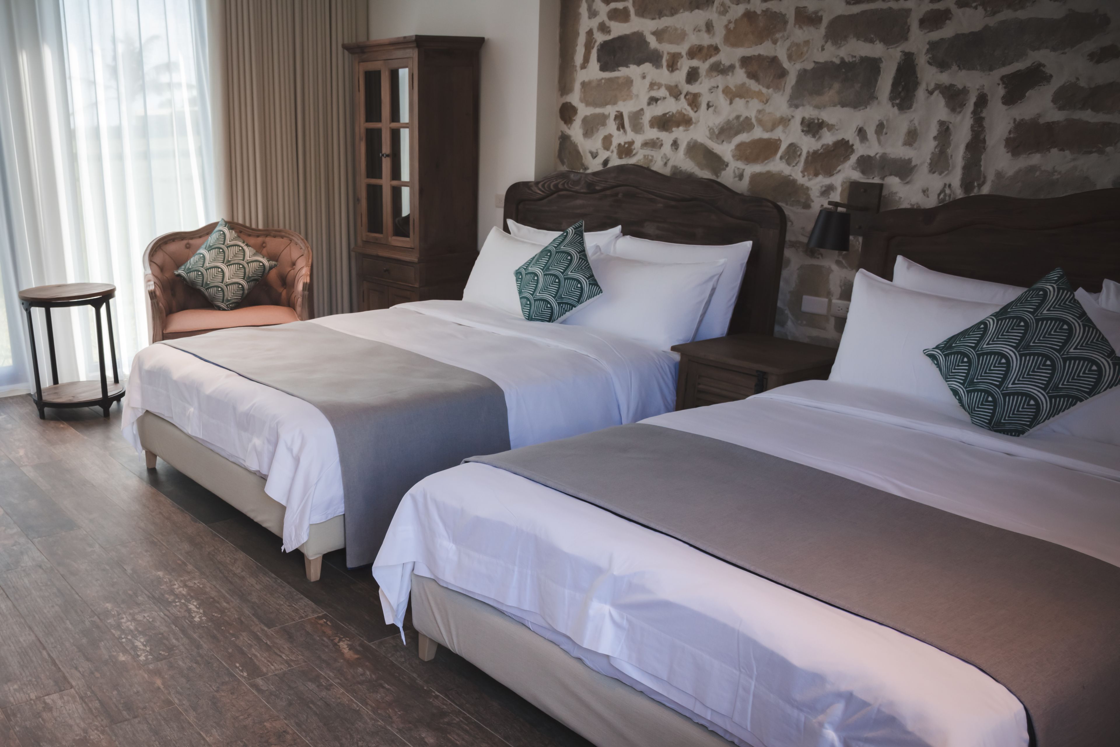Two beds with white linens, a wooden chair, a small table, and a stone wall.