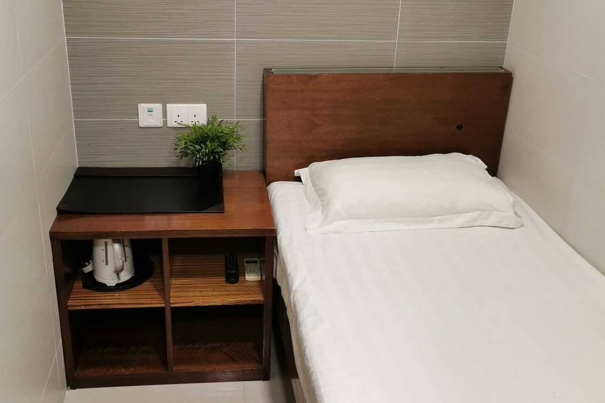 A small, neatly arranged bedroom with a wooden nightstand, a single bed with white bedding, and a small shelf with a plant and a white bag.