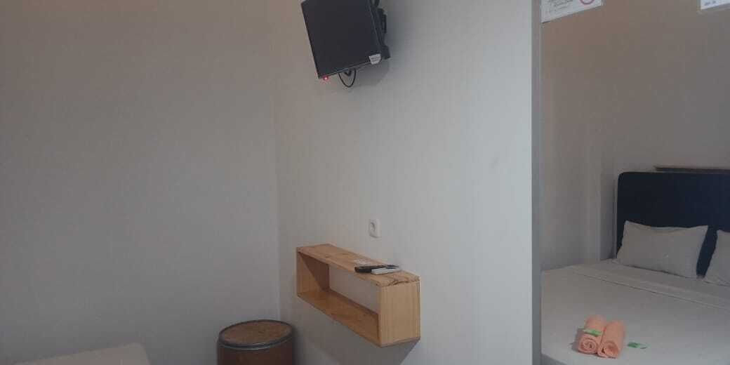 A small, simple room with a bed, a wooden shelf, a trash can, and a wall-mounted TV.