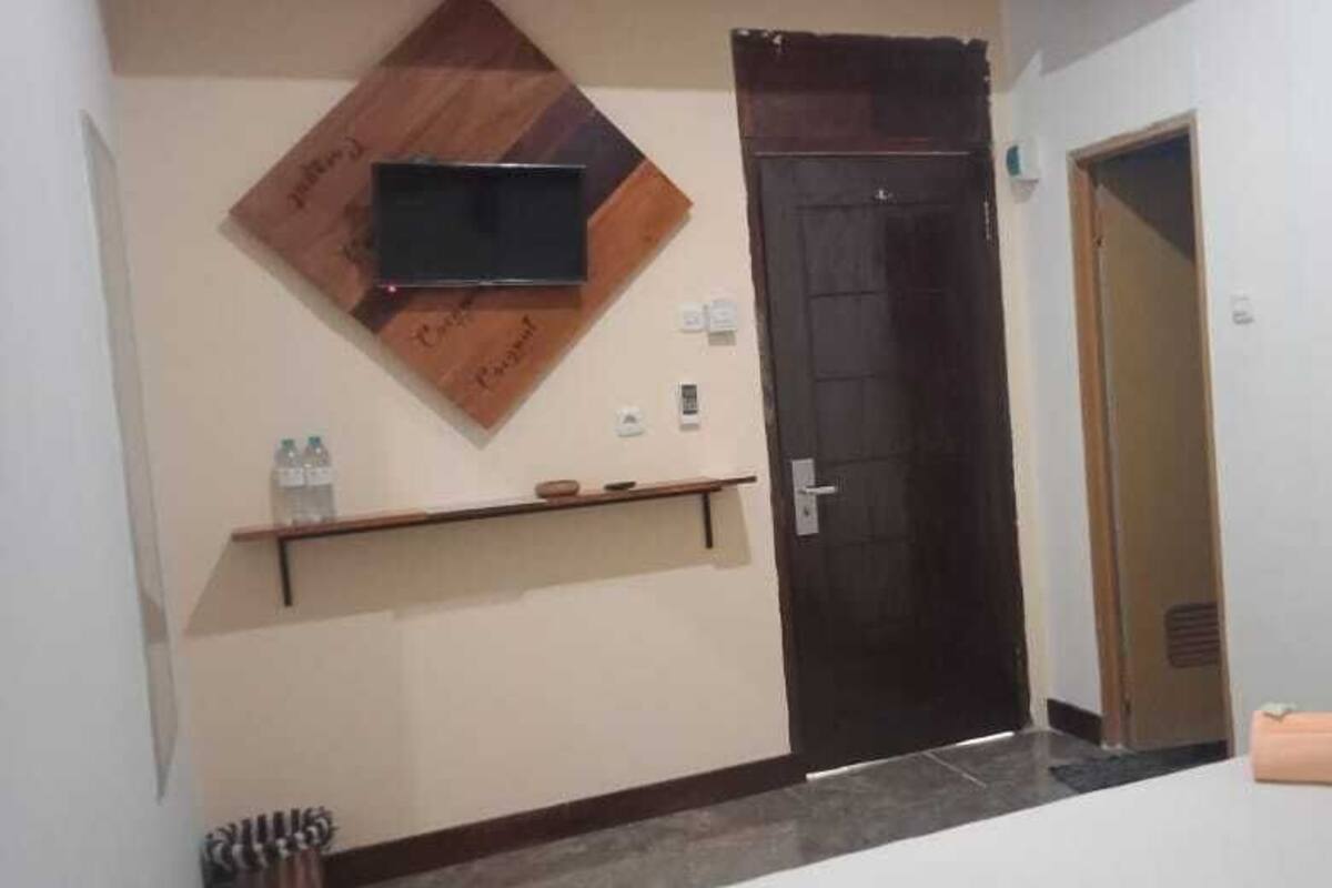 A hotel room with a dark wooden door, a mounted TV, a small shelf with a water bottle, and a trash bin.