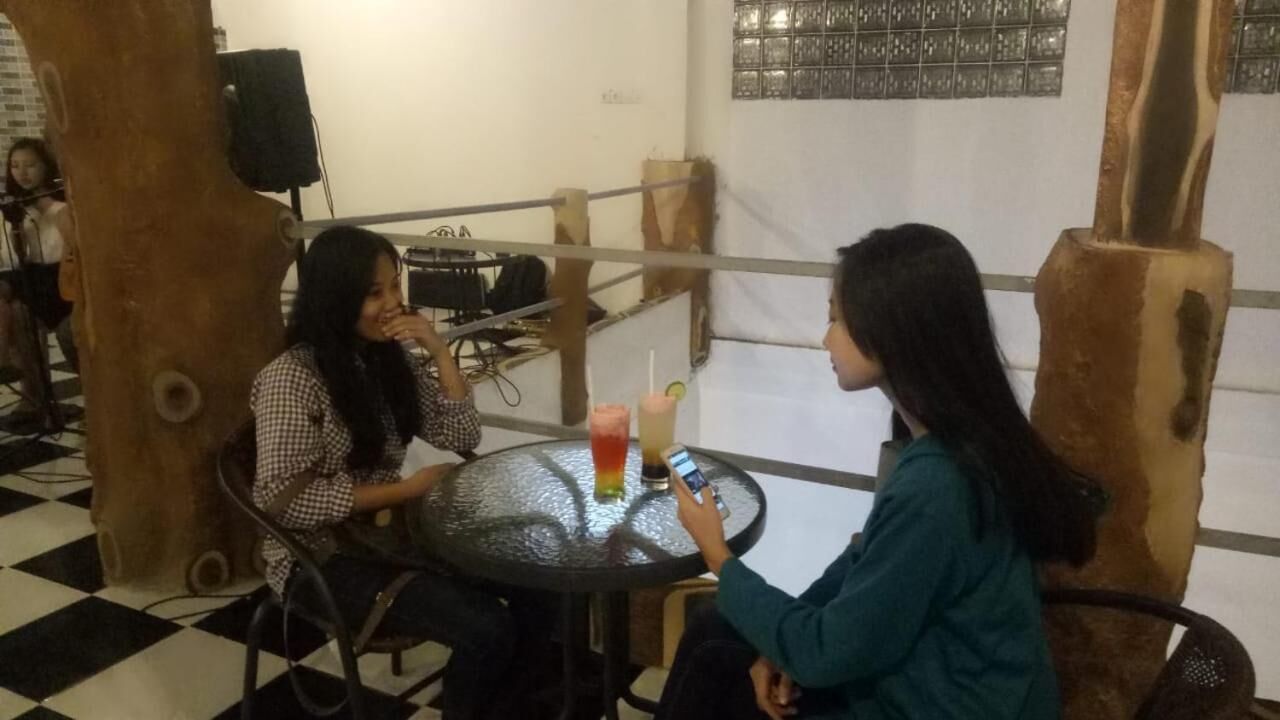 Duas pessoas sentadas numa mesa redonda com bebidas, num espaço com chão quadriculado e pilares de madeira.