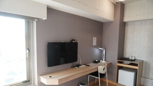 A modern hotel room with a wooden desk, a flat-screen TV, a desk lamp, a chair, a small refrigerator, and a trash can. - Jia Inn (Tainan)