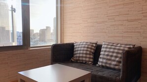A modern living room with a sofa and a small table, a large window with a city view, and a brick-patterned wall. - Jia Inn (Tainan)