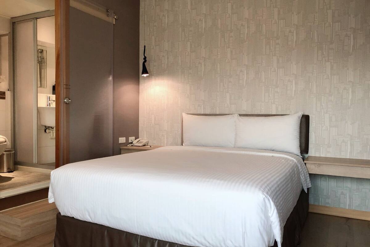 Una habitación de hotel con una cama grande, una mesita de noche con lámpara y un papel tapiz con vista a la ciudad.