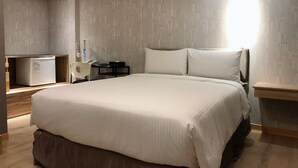 A modern hotel room with a large bed, a bedside table, a desk with a chair, a small refrigerator, and a wall-mounted lamp. - Jia Inn (Tainan)