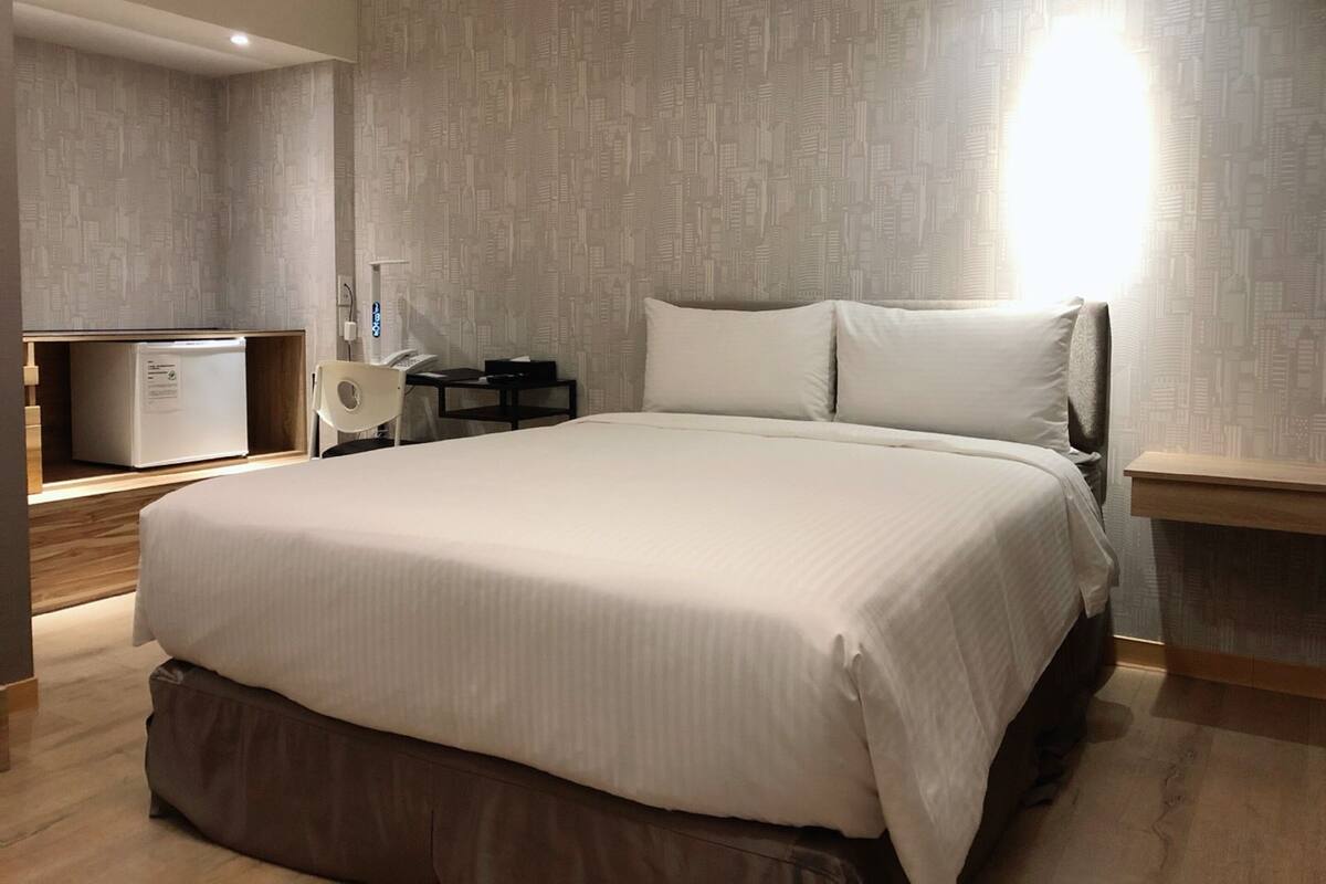 Una habitación de hotel moderna con una cama grande, una mesita de noche, un escritorio con una silla, un refrigerador pequeño y una lámpara fijada a la pared.